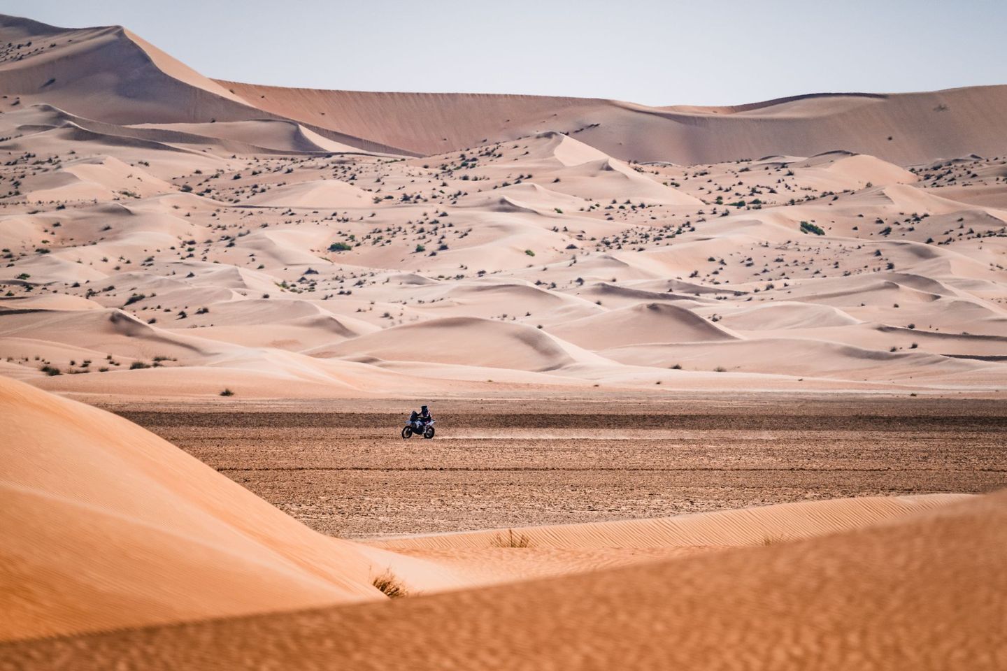 Alex McInnes riding his rally bike across a flat between towering sand dunes