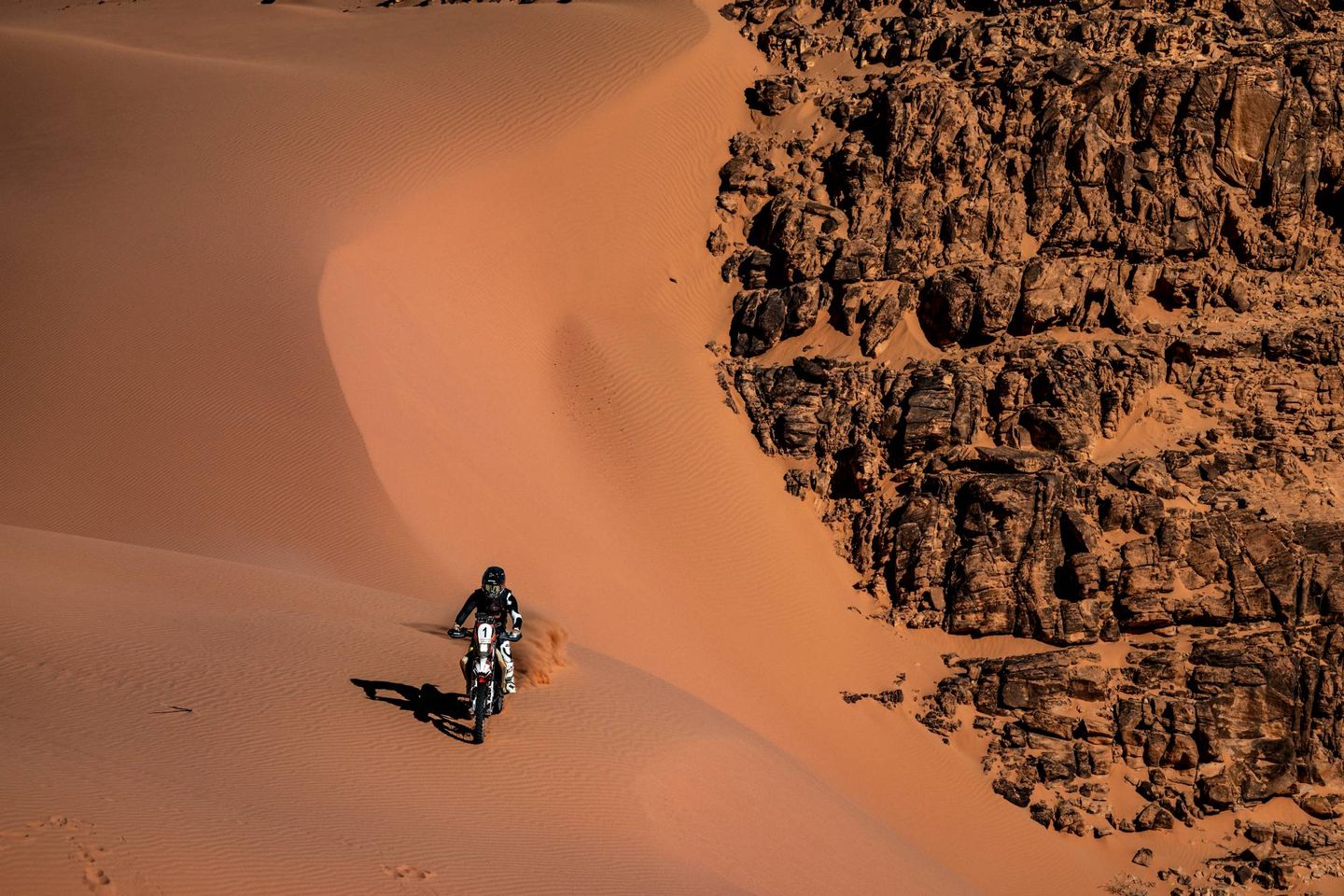 Alex McInnes riding down a red dune beside a wall of rock